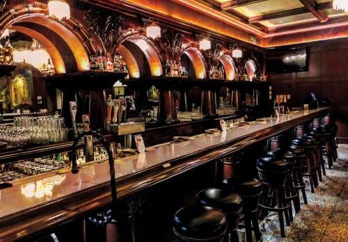 A dimly lit bar with a long counter, ornate wooden details, and shelves lined with bottles. Stools are arranged along the bar.