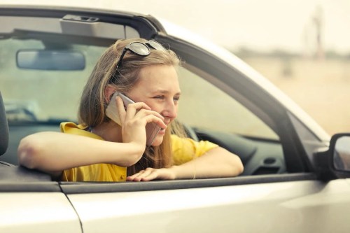 A woman in a yellow shirt leans out of a car window, smiling while talking on her phone.