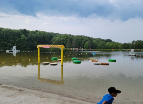 A boy runs along the shore of a lake with colorful floating rings and a yellow inflatable arch in the background.
