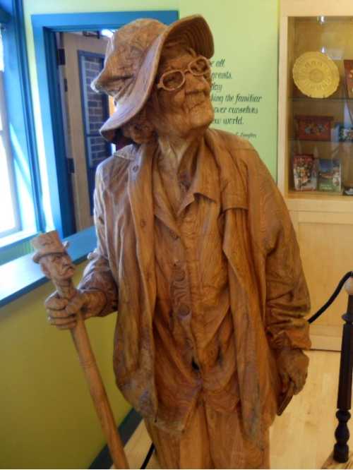 Wooden sculpture of an elderly woman in a hat and glasses, holding a walking stick, set in a colorful room.