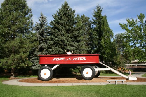 A large red Radio Flyer wagon sits on a grassy area surrounded by trees under a blue sky.