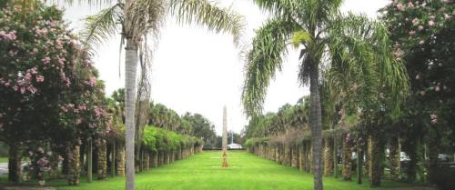 A lush green pathway lined with palm trees and flowering plants, leading to a central sculpture in a serene landscape.