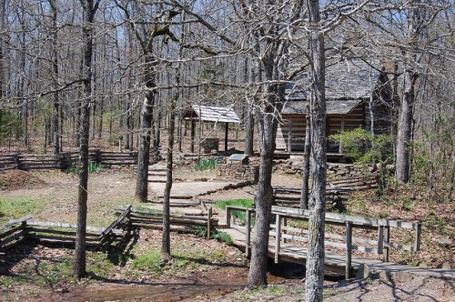 A rustic cabin surrounded by trees, with a stone path and wooden bridge leading to it in a serene forest setting.