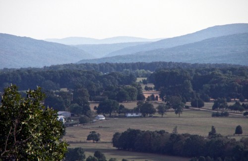 A scenic view of rolling hills and farmland under a hazy sky, with trees and distant mountains in the background.