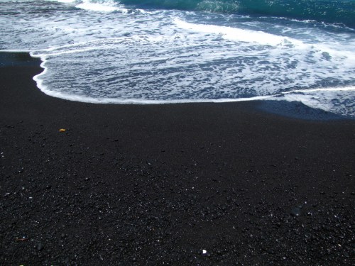 Waves gently lap at a black sand beach, creating a contrast with the white foam of the water.