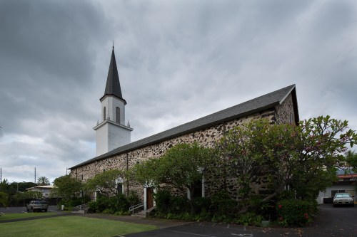 A stone church with a tall spire, surrounded by greenery, under a cloudy sky.