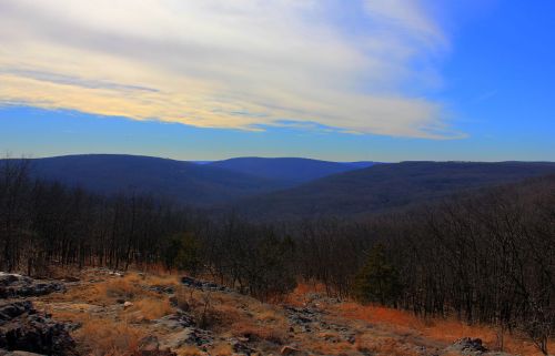 A panoramic view of rolling hills and valleys under a blue sky with wispy clouds, surrounded by sparse trees.