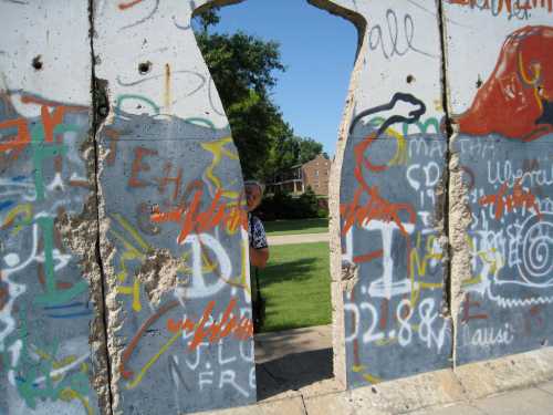 A person peeks through a colorful, graffiti-covered section of the Berlin Wall in a green park setting.