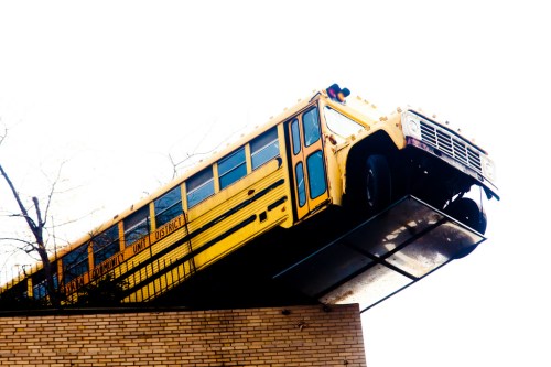 A yellow school bus is precariously perched on the edge of a building, with its front wheels hanging off.