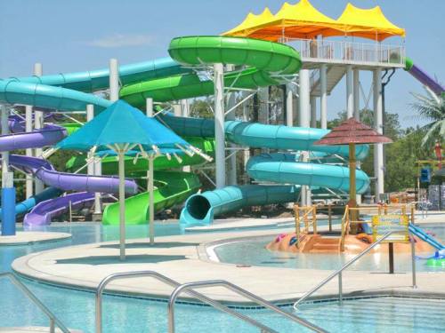 Colorful water slides and a splash area by a pool, with bright umbrellas and a sunny sky in the background.