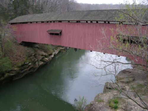 A red covered bridge spans a green river, surrounded by rocky banks and bare trees in a serene landscape.