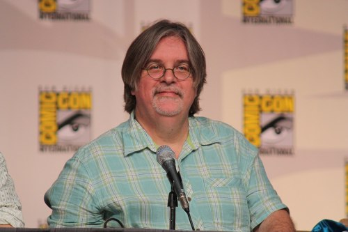 A man with glasses and long hair smiles at a microphone, with a Comic-Con backdrop behind him.