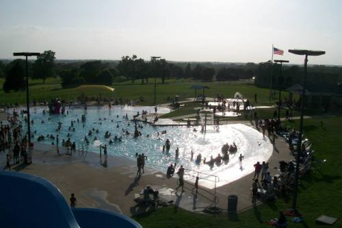 A busy outdoor pool with people swimming and playing, surrounded by green grass and a clear sky. An American flag is visible.