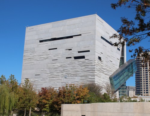 Modern building with a textured gray facade and angular glass elements, surrounded by trees and a clear blue sky.