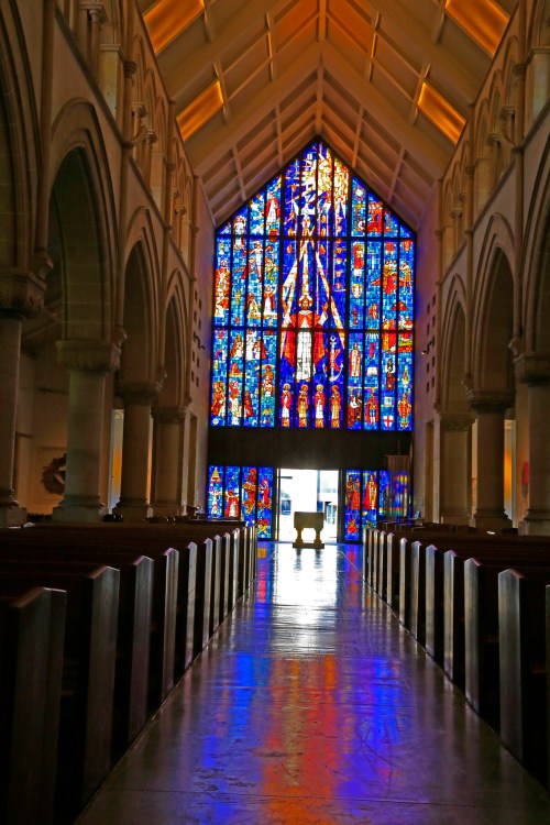 A church interior featuring a tall stained glass window, colorful light reflecting on the polished floor.