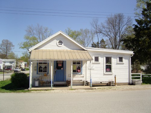 A small white house with a blue door and a metal roof, surrounded by trees and a gravel path.