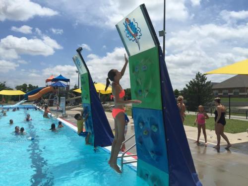 A girl in a pink swimsuit jumps towards a climbing wall at a sunny pool, with children playing in the water nearby.