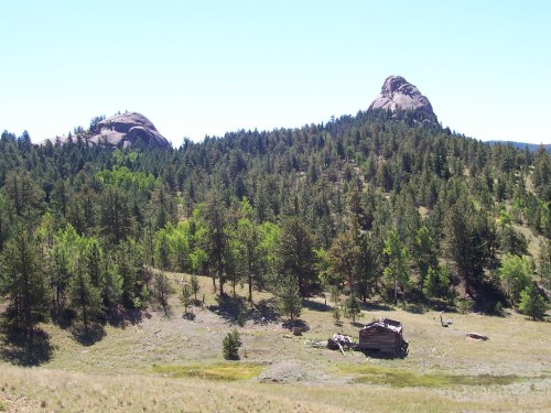 A scenic landscape featuring rocky hills, lush green trees, and a small, weathered cabin in a grassy area.