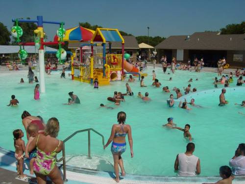 A busy water park with children playing in a pool, colorful slides, and a play structure under a clear blue sky.