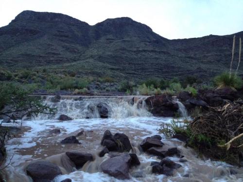 A rocky stream cascades over boulders, with a mountainous landscape in the background under a cloudy sky.