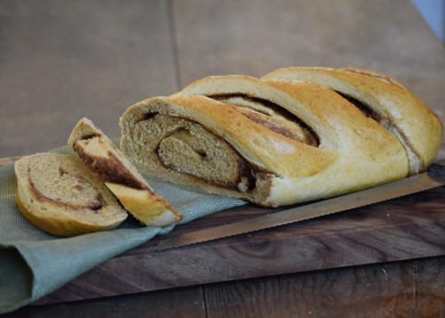 A freshly baked braided loaf of cinnamon bread, with a slice cut and resting beside it on a wooden board.