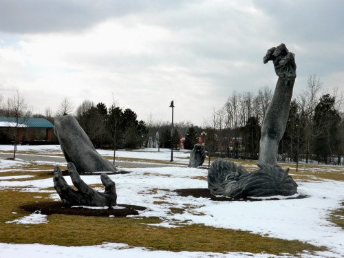 A park scene featuring large, abstract sculptures of hands and arms emerging from the ground, surrounded by snow.