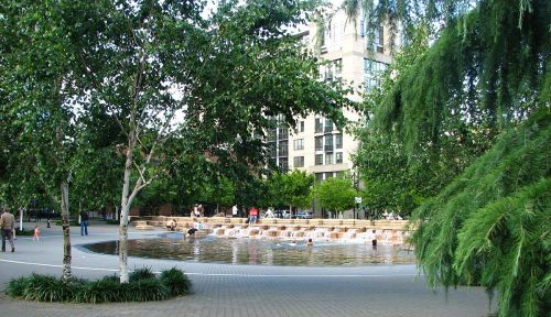 A public park with a shallow fountain, surrounded by trees and people enjoying the water on a sunny day.