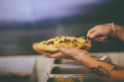 A person in gloves holds a loaded hot dog topped with cheese and meat, ready to be served.