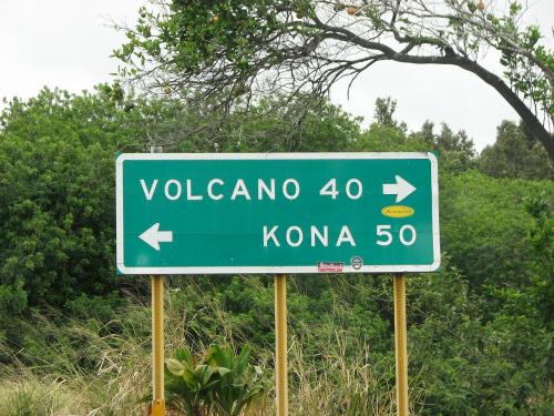 Green road sign indicating directions to Volcano (40 miles) and Kona (50 miles), surrounded by greenery.