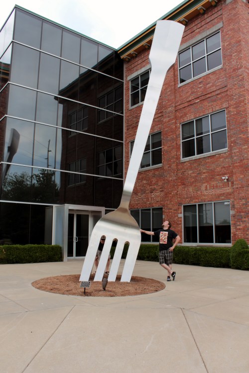 A giant silver fork sculpture stands outside a modern brick building, with a person posing next to it.