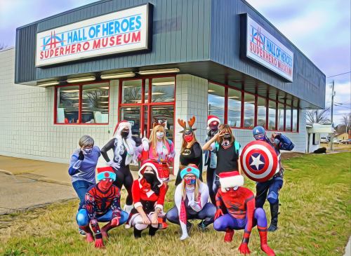 A group of costumed superheroes poses outside the Hall of Heroes Superhero Museum, showcasing festive attire.