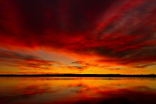 Vibrant sunset over a calm lake, with dramatic red and orange clouds reflecting on the water's surface.