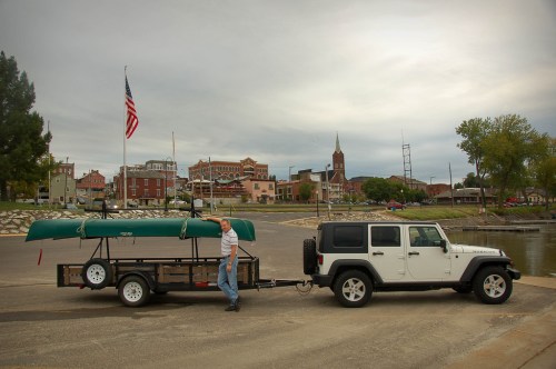 A man pulls a trailer with a canoe behind a white Jeep at a waterfront, with a small town and cloudy sky in the background.