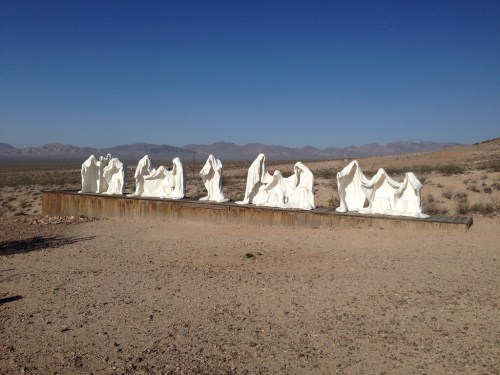 A row of white sculptures on a desert landscape, depicting abstract figures against a clear blue sky.