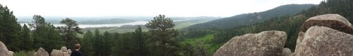 A panoramic view of a lush green landscape with hills, a lake, and a person standing among large rocks.