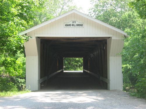 A covered bridge with a sign reading "1872 Adams Mill Bridge," surrounded by greenery.