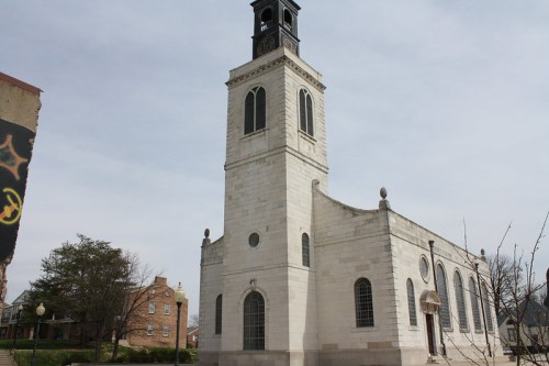 Historic church with a tall clock tower, featuring large windows and a stone facade, set against a cloudy sky.