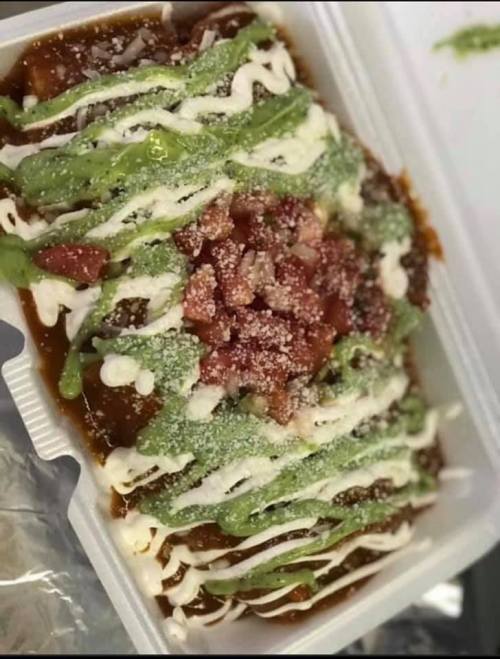 A takeout container filled with layered enchiladas topped with green sauce, sour cream, diced tomatoes, and cheese.