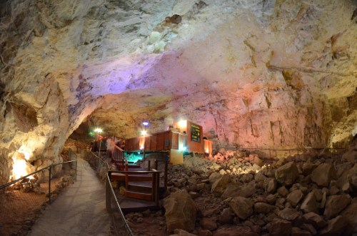 A spacious underground cave with colorful lighting, rocky terrain, and wooden furniture set up for visitors.