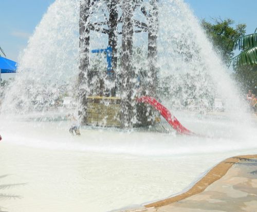 A water play area with a large fountain, a red slide, and people enjoying the splash zone on a sunny day.