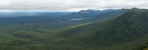 A panoramic view of lush green mountains and valleys under a cloudy sky, with a lake visible in the distance.