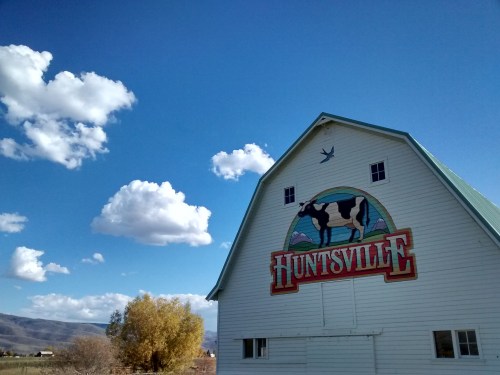 A white barn with a colorful "Huntsville" sign and a cow mural, set against a blue sky with fluffy clouds.