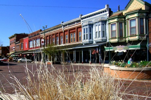 Historic buildings line a sunny street, with dry grass in the foreground and a clear blue sky above.
