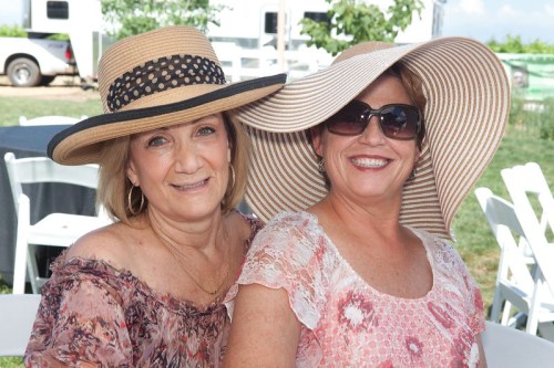 Two women wearing large sun hats and sunglasses smile together at an outdoor event.