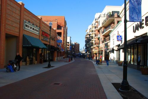 A wide, sunny street lined with shops and apartments, featuring pedestrians and outdoor seating.