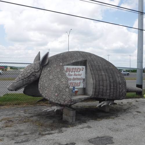 A large, concrete armadillo sculpture with a sign for "Bussey's Flea Market" in Texas, set against a cloudy sky.