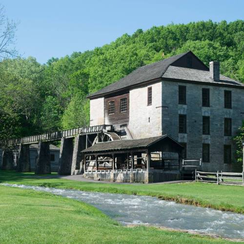 Historic stone mill beside a flowing stream, surrounded by lush green trees and a clear blue sky.