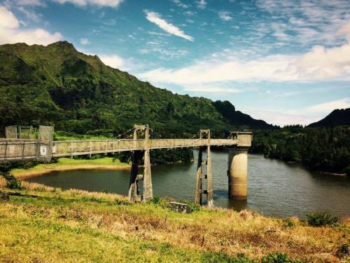 A scenic view of a bridge over a river, surrounded by lush green mountains and a blue sky with scattered clouds.