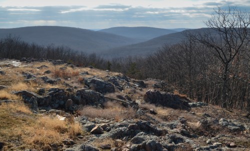 A rocky landscape with sparse vegetation, overlooking a valley and distant mountains under a cloudy sky.