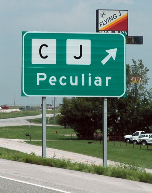 Green highway sign indicating direction to Peculiar, with symbols for CJ and an arrow pointing right.